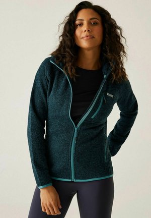 Kobieta z kręconymi włosami, ubrana w ciemny turkusowy polar zapinany na suwak, na czarnej bluzce, stojąca z jedną ręką na biodrze na jednolitym tle.