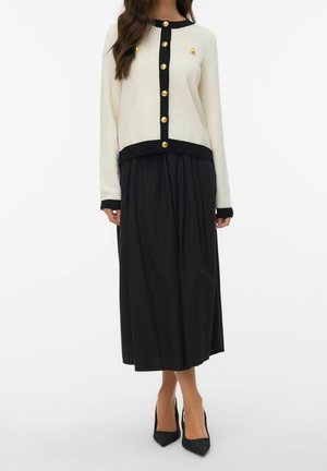 Femme portant un cardigan crème avec bordure noire et boutons dorés, associé à une jupe midi plissée noire et des talons noirs pointus.
