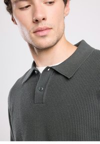 Tumma vihreä poolopaita, jossa on teksturoitu neulos, klassinen kaulus ja kaksi mustalla napilla varustettua kiinnitystä.