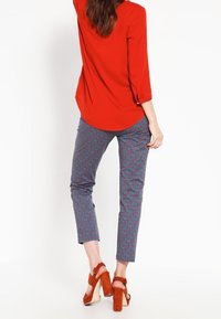 Červená blůza s tříčtvrtečními rukávy v kombinaci s modrými vzorovanými kalhotami. Červené lodičky na vysokém podpatku s kotníkovými pásky dotvářejí celý outfit.