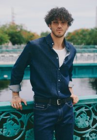 Camisa de mezclilla en azul oscuro con puños en azul claro, combinada con jeans azul oscuro. Cinturón negro con hebilla plateada. El fondo presenta una barandilla turquesa.