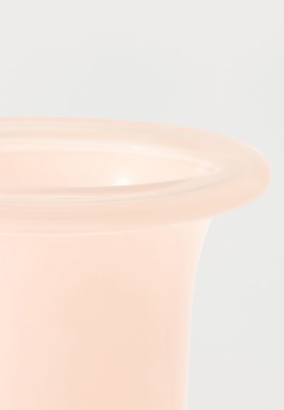 Κοντινό πλάνο ενός διαφανούς γυάλινου βάζου σε ανοιχτή ροδακινί απόχρωση, με στρογγυλεμένο, φουσκωτό χείλος, πάνω σε απλό φωτεινό φόντο.