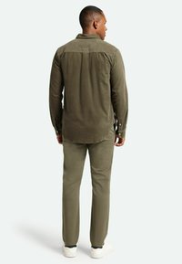Camicia verde oliva con colletto button-down, maniche lunghe e orlo leggermente curvo, abbinata a pantaloni verde oliva coordinati e sneaker bianche.