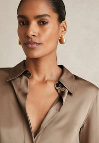 Femme aux cheveux lisses tirés en arrière, portant des boucles d'oreilles pendantes en or et un chemisier en satin taupe avec un col ouvert sur un fond neutre.