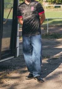 Maglia nera con un motivo testurizzato e dettagli rosa, abbinata a jeans blu larghi e scarpe nere, in piedi vicino a un autobus.