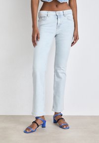 Jeans de mezclilla azul claro con un corte ajustado, que presentan una cintura de altura media y un ligero desgaste. Combinados con sandalias de tacón con tiras en negro y azul.
