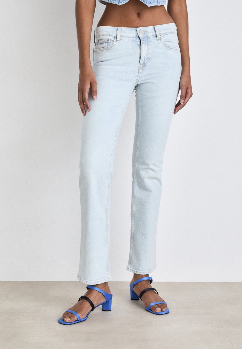 Jeans de mezclilla azul claro con un corte ajustado, que presentan una cintura de altura media y un ligero desgaste. Combinados con sandalias de tacón con tiras en negro y azul.