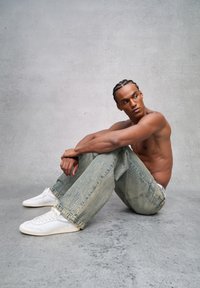 Jeans de mezclilla de lavado claro con un ajuste relajado, que presentan dobladillos deshilachados y detalles bordados. Combinados con zapatillas blancas. Pose casual.