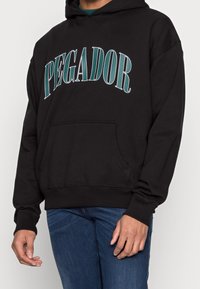 Felpa nera con un grande design del testo "PEGADOR" in teal. Presenta una tasca a marsupio e polsini a costine. Realizzata in tessuto morbido.