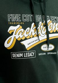 Sweat à capuche vert foncé avec texte blanc et jaune mentionnant « Fine Cotton Select, Trademark, Jack & Jones, Denim Legacy, Barcelona, Copenhagen. »