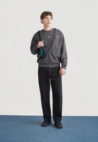 Γκρι φούτερ με φερμουάρ και πλαϊνές τσέπες, συνδυασμένο με μαύρο παντελόνι και μαύρα παπούτσια. Μικρά λογότυπα είναι ορατά στα ρούχα.