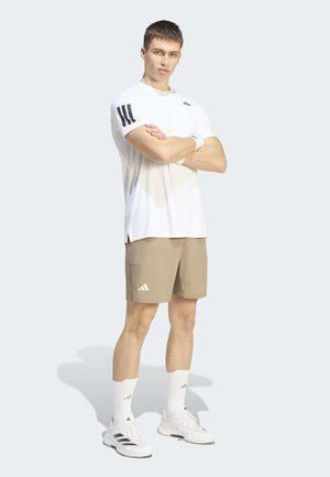 Junger Mann steht mit verschränkten Armen, trägt weißes T-Shirt, beige Shorts, weiße Socken und weiße Adidas-Sneakers auf hellem Hintergrund.