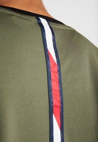 Olivgrön t-shirt med nätstruktur, med en vertikal randdesign i marinblått, vitt och rött längs axelsömmen.