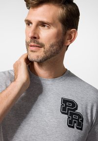 Szara bawełniana koszulka z krótkim rękawem, z okrągłym dekoltem i czarnym logo "PR" na lewej piersi. Materiał wydaje się gładki.
