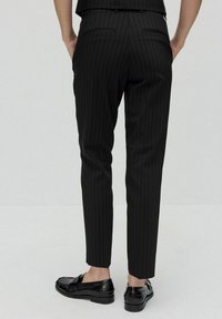 Svarta byxor med nålrand, i en figurnära passform, med bakfickor. Kombinerade med svarta loafers, mot en neutral bakgrund.