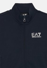 Námornícka bunda na zips so stojatým golierom, s strieborným logom "EA7 Emporio Armani" na ľavej strane a hladkou textúrou látky.