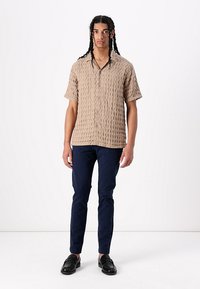 Beige kortærmet skjorte med tekstureret rutenetmønster, knaplukning foran, båret med marineblå bukser og sorte sko. Enkel, afslappet design.