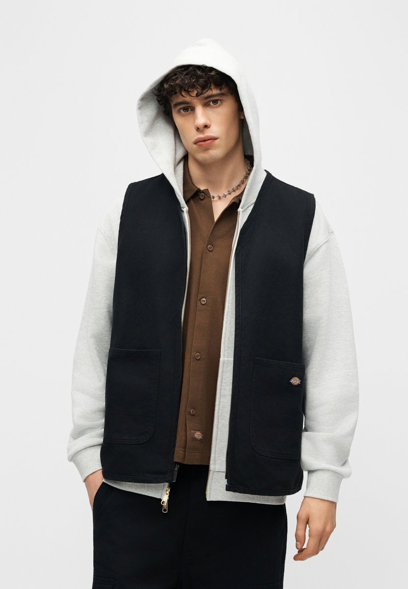 Giovane uomo che indossa una felpa grigia con cappuccio sotto un gilet nero senza maniche e una camicia marrone con bottoni, in piedi davanti a uno sfondo semplice.