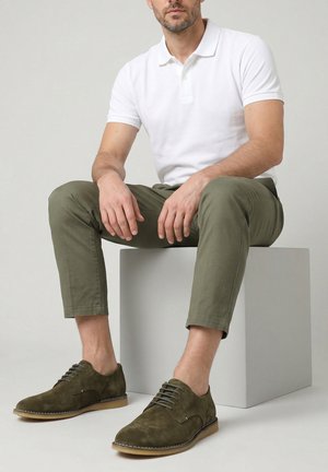 Man zit op een witte kubus, draagt een wit poloshirt met korte mouwen, olijfgroene broek die boven de enkels is afgeknipt en olijfgroene suède veterschoenen.