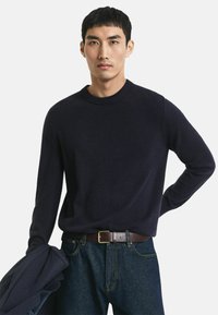 Marineblauer Rundhals-Pullover aus weichem Stoff mit langen Ärmeln, getragen mit einem braunen Gürtel und dunklen Jeans, gehalten von einer marineblauen Jacke.