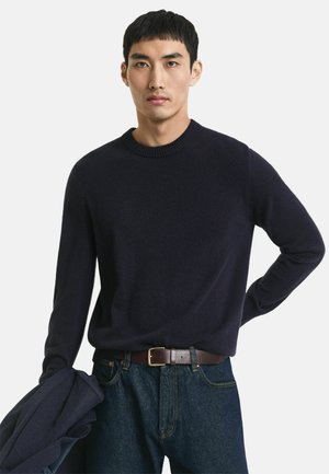 Marineblauer Rundhals-Pullover aus weichem Stoff mit langen Ärmeln, getragen mit einem braunen Gürtel und dunklen Jeans, gehalten von einer marineblauen Jacke.