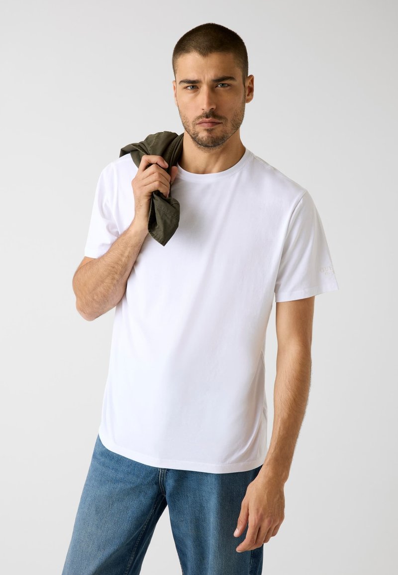 Weißes Baumwoll-T-Shirt mit Rundhalsausschnitt, kurzen Ärmeln und lockerer Passform. Kombiniert mit blauen Denim-Jeans, hält eine dunkelgrüne Jacke.