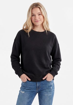 Ung kvinde med langt blondt hår iført en sort sweatshirt og revnede blå jeans, stående med hænderne i lommerne mod en hvid baggrund.