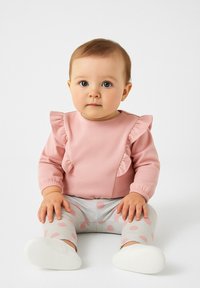 Ružová blúzka s dlhým rukávom a volánovými detailmi, v kombinácii so šedými legínami s ružovými bodkami. Oblečenie je vyrobené z jemnej, štruktúrovanej látky.