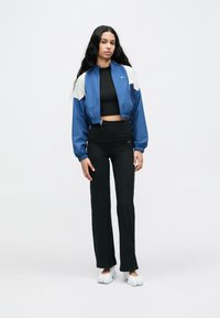 Μπλε και λευκό αδιάβροχο με ελαστικά μανσέτα, φορεμένο πάνω από μαύρο κοντό τοπ και μαύρο παντελόνι ψηλόμεσο. Οι λευκές αθλητικές παπούτσια ολοκληρώνουν το σύνολο.