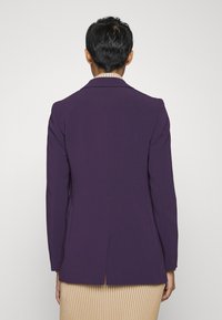 Fioletowa marynarka o dopasowanym kroju, z pojedynczym rozcięciem z tyłu i zbudowanymi ramionami. Nosi się ją na beżowej spódnicy w paski.