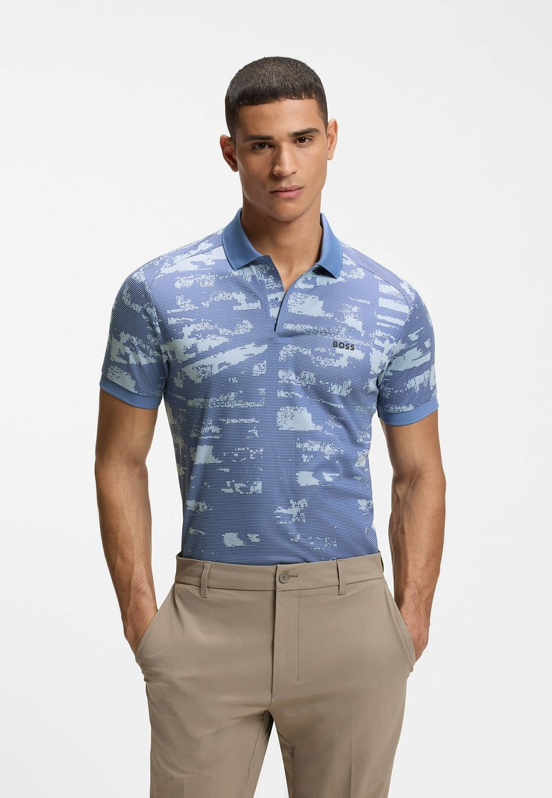 Homme portant un polo bleu à motifs avec le logo "BOSS" et un pantalon beige, debout les mains dans les poches sur un fond blanc.