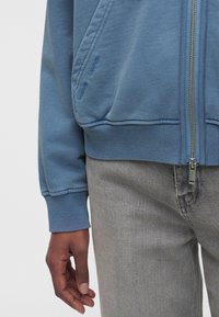 Gros plan d'une personne portant une veste bleue à zip avec des poignets côtelés et un jean gris, montrant une main détendue le long du corps.