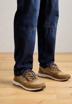 Sneakers marroni e beige con lacci indossati con jeans blu scuro, in piedi su un pavimento di legno.