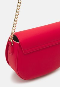 Červená kabelka z umělé kůže s oblým tvarem, hladk�ým povrchem a zlatým řetězovým popruhem. Obsahuje klopu a minimální detaily šití.
