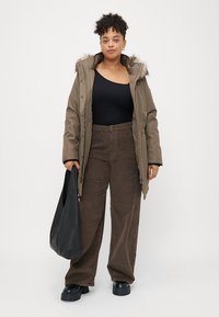 Casaco acolchoado castanho com capuz forrado a peluche, blusa preta com texturização, e calças de veludo cotelê castanhas. Modelo segura uma mala de ombro preta, de pé contra um fundo neutro.