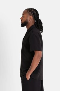 Zwart shirt met korte mouwen, kraag, pocketdetail en een gladde textuur, gedragen met donkere onderkleding. Model staat in profiel.