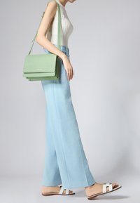 Borsa a tracolla verde con texture e design a strati, hardware dorato, indossata con pantaloni a gamba larga blu chiaro e sandali bianchi, che mostrano un look minimalista.