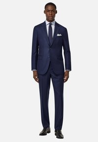Costume sur mesure bleu marine avec une veste à un bouton, des revers crantés et un pantalon assorti. Comprend une chemise bleu clair et une cravate sombre.