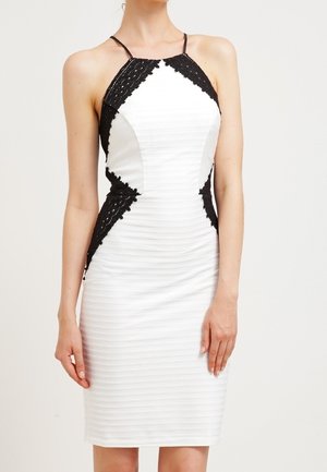 Biała dopasowana sukienka bez rękawów z czarnymi koronkowymi ozdobami po bokach, z wysokim dekoltem i cienkimi czarnymi ramiączkami, noszona przez stojącą osobę.