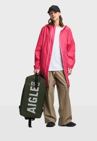 Personne portant un imperméable rose, une chemise blanche, un pantalon beige, des chaussures noires, une casquette noire, tenant un sac vert de la marque Aigle à ses côtés.