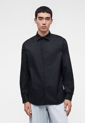 Jeune homme portant une chemise noire à manches longues boutonnée et un pantalon gris clair, debout devant un fond blanc uni.