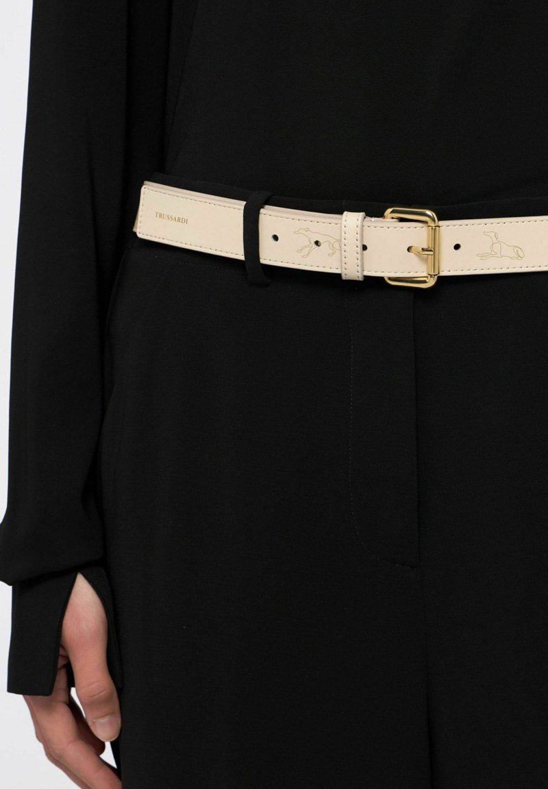 Beige leren riem met goudkleurige gesp, gestikte details en een subtiele logo-opdruk, gedragen over een zwart kledingstuk.