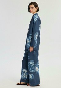 Traje de tie-dye azul que presenta una chaqueta larga y pantalones de pierna ancha, hecho de una tela parecida al denim con una textura suave. Incluye pendientes con acentos dorados.