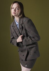 Blazer marrón de espiga con diseño de doble botonadura, botones negros y mangas largas, combinado con una falda mini texturizada y una blusa azul clara.