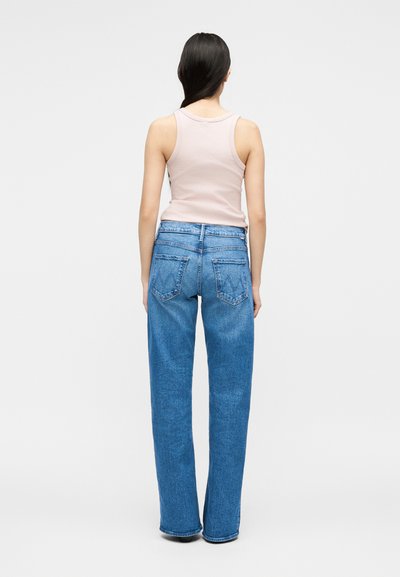 Γυναίκα που φαίνεται από πίσω με ροζ αμάνικη μπλούζα ανοιχτού χρώματος και μπλε τζιν ψηλόμεσα, με ίσια μπατζάκια, σε απλό φωτεινό φόντο.