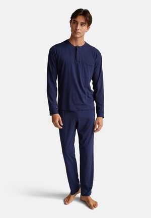 Junger Mann, der barfuß in einem dunkelblauen Langarm-Henley-Shirt und passenden Hosen vor einem schlichten weißen Hintergrund steht.