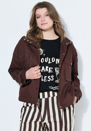 Młoda kobieta w brązowej kurtce, czarnej koszulce z nadrukiem, spodniach w paski i złotych kolczykach kołach, stojąca na tle białego tła.