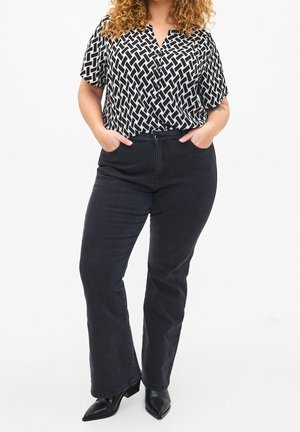 Femme aux cheveux bouclés portant un chemisier à motif noir et blanc, un jean noir taille haute à jambes larges et des chaussures noires pointues, les mains dans les poches.