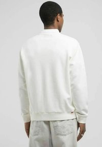 Felpa bianca con collo a giro, polsini e orlo a costine, tessuto di peso medio, design minimalista, vista posteriore con etichetta logo vicino all'orlo.