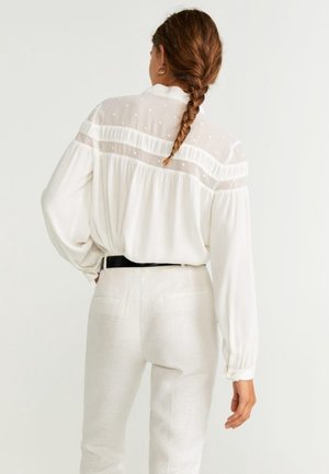 Blusa bianca con retro trasparente a pois, dotata di maniche lunghe e dettagli arricciati. Abbinata a pantaloni chiari e una cintura scura.